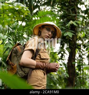 Donna botanica vestita in stile safari in serra. Naturalista in abiti kaki, guanti di lievito con zaino passeggiate nella foresta pluviale circondata da p. Foto Stock