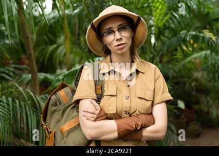 Primo piano ritratto di donna botanica vestita in stile safari in serra. Naturalista in abiti cachi, guanti di lievito con zaino guardando la macchina fotografica, Foto Stock