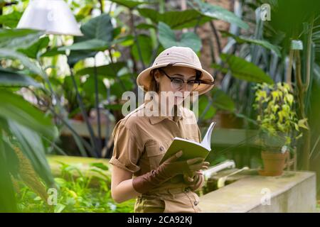 Donna botanica vestita in stile safari in serra. Naturalista che indossa cappello e abiti cachi, guantoni di lievito che riposano e leggendo un libro circondato da Foto Stock