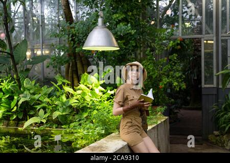 Donna botanica vestita in stile safari in serra. Naturalista che indossa cappello e abiti cachi, guantoni di lievito che riposano e leggendo un libro circondato da Foto Stock