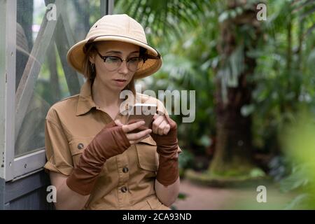 Donna botanica vestita in stile safari in serra. Naturalista in abiti cachi, guantoni di lievito che riposano e leggono un libro, indoor. Foto Stock