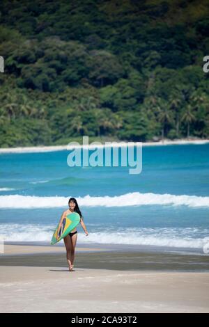 Beach shot di un brasiliano giapponese (Nipo-brasileiro) in un bikini che porta una tavola da surf decorata con la bandiera brasiliana, Brasile, Sud America Foto Stock