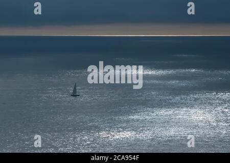 Barca a vela da sola nell'oceano Pacifico vicino alla costa del Cile. Il sole all'orizzonte crea con il cielo nuvoloso riflessi di luce sul se Foto Stock