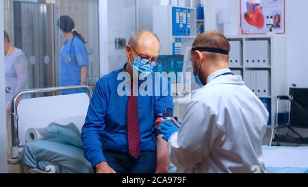 Rack Focus dall'ascensore di un ospedale occupato o clinica privata, semeborpo indossare maschere. Medico che misura la pressione sanguigna, infermiera che aiuta la vecchia signora in pensione. Tema COVID-19. Medicina medica sistema sanitario Foto Stock