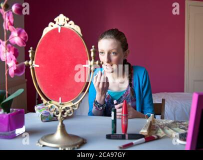 Giovane ragazzina di quattordici anni si siede nella sua camera da letto applicando trucco in uno specchio in preparazione per uscire. Foto Stock