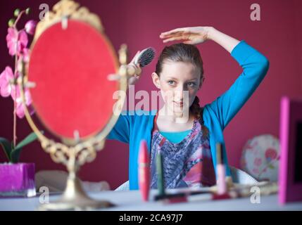 Quattordici anni ragazza teenage si siede nella sua camera da letto applicando trucco in uno specchio sulla sua tabella di vestizione in preparazione di uscire. Foto Stock