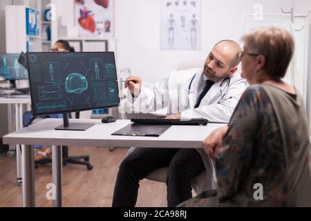 Medico che indica la scansione tc sul computer per i pazienti anziani durante la visita dopo l'intervento chirurgico cerebrale. Diagnosi per trauma cranico. Medico indossando camice bianco e stetoscopio. Foto Stock