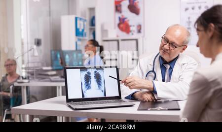 Medico senior che punta alla radiografia polmonare su un computer portatile per i pazienti giovani in ospedale. Donna disabile matura in sedia a rotelle che discute con medic circa la malattia. Foto Stock