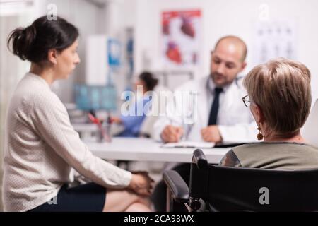 Donna anziana disabile in sedia a rotelle con figlia che ascolta medico durante la consultazione in ospedale ufficio. Medico indossando camice bianco e stetoscopio prendendo appunti durante l'esame. Foto Stock