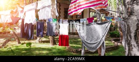 Scena di vita reale domestica di molti bambini e adulti vestiti puliti lavati appesi su stendibiancheria di betulla con spille. Cortile di casa su luminoso sole Foto Stock