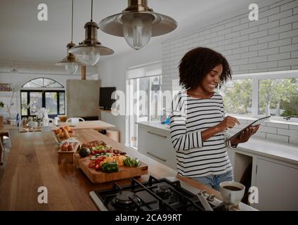 Giovane allegra donna afroamericana che controlla la ricetta su una tavoletta digitale mentre prepara l'insalata di verdure in cucina Foto Stock