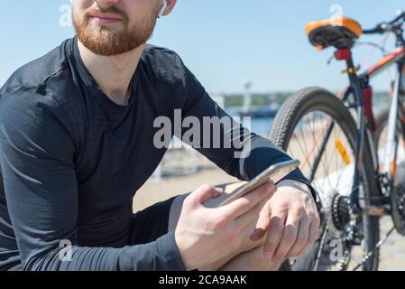 Ritratto di un atleta di ciclista che riposa dopo un giro in bicicletta usando uno smartphone in mano. All'aperto. Sport primaverili. Il ragazzo è venticinque voi Foto Stock