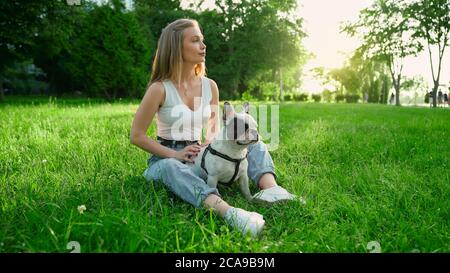 Vista laterale della giovane donna felice seduta su erba fresca con carino bulldog bianco e marrone francese. Splendida ragazza sorridente godendo il tramonto estivo, cane accarezzante nel parco cittadino. Amicizia umana e animale. Foto Stock