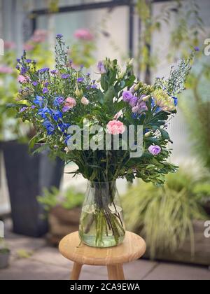 Bel bouquet grande di fiori in vaso esterno esposto su un piccolo tavolo di legno. Plantlife fioritura in piedi in acqua. Sfondo sfocato, garofani blu-viola e bianco foglie verdi. Foto Stock