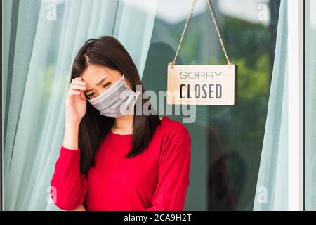 Asian giovane donna indossare maschera viso proteggere Sad avviso segno etichetta tavola di legno 'SORRY SIAMO chiusi SI PREGA DI TORNARE INDIETRO' appeso attraverso porta di vetro Foto Stock