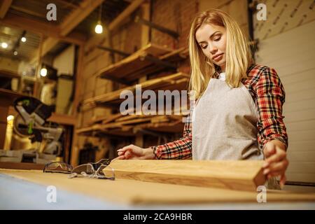 giovane carpentiere caucasica professionale donna controllo per ruvidità pezzo di legno dopo la macinazione, femmina in lavoro uniforme in fabbrica Foto Stock
