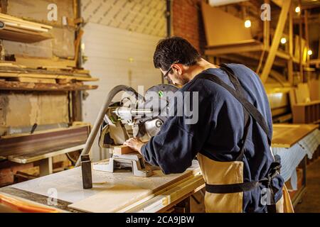 giovane falegname caucasico uomo al lavoro, bel ragazzo in abiti da lavoro utilizzare trapani elettrici e altre macchine per il taglio del legno Foto Stock