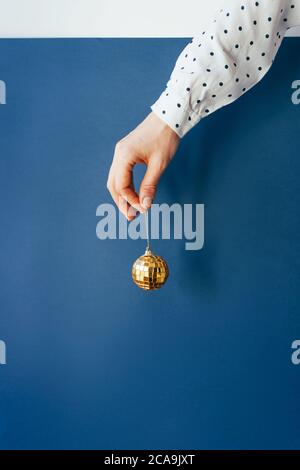 La mano di una donna in una camicia a pois tiene una pallina di Natale dorata su sfondo blu. Foto Stock