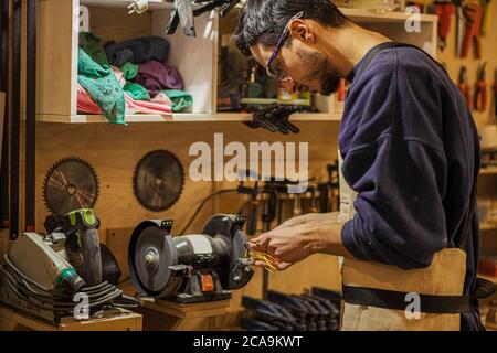 giovane falegname duro che lavora segando e tagliando legno sulla macchina nel suo studio, indossando occhiali protettivi sugli occhi e uniforme speciale Foto Stock