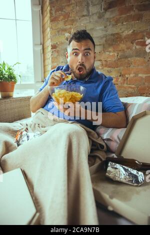 Scioccati, che mordono i critici. Uomo pigro che vive nel suo letto circondato da sudicio. Non c'è bisogno di uscire per essere felici. L'utilizzo di gadget, la visione di film e serie, sembra emotivo. Mangiare spuntini e fast food. Foto Stock