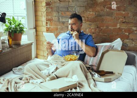 Scioccati, che mordono i critici. Uomo pigro che vive nel suo letto circondato da sudicio. Non c'è bisogno di uscire per essere felici. L'utilizzo di gadget, la visione di film e serie, sembra emotivo. Mangiare spuntini e fast food. Foto Stock