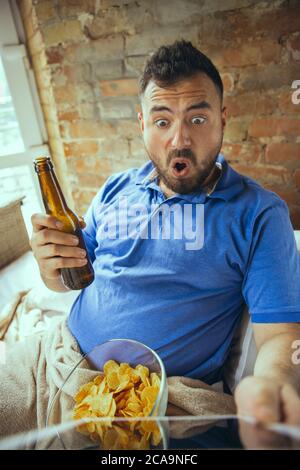 Scioccato, beve birra. Uomo pigro che vive nel suo letto circondato da sudicio. Non c'è bisogno di uscire per essere felici. L'utilizzo di gadget, la visione di film e serie, sembra emotivo. Mangiare spuntini e fast food. Foto Stock