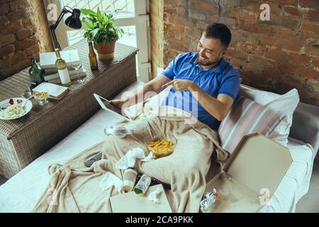 Sorridendo, puntando durante la videocall utilizzando il tablet. Uomo pigro che vive nel suo letto circondato da sudicio. Non c'è bisogno di uscire per essere felici. Utilizzo di gadget, guardare film e serie, emotivo. Fast food. Foto Stock