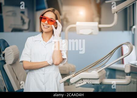 bella donna di buon aspetto che lavora come dentista in clinica indossando uniforme bianca e occhiali rossi per la sicurezza, in posa in studio dentistico Foto Stock