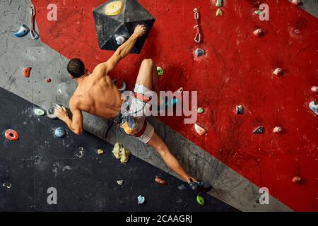 Panoramica del giovane uomo muscolare in allenamento indoor clymbing palestra. Vista posteriore di uno sportivo scalatore nello spostamento verso l'alto sulla roccia ripida, arrampicata su w artificiale Foto Stock