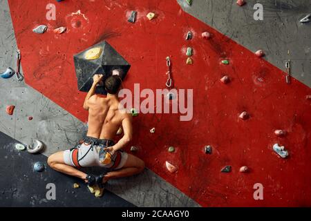 Panoramica del giovane uomo muscolare in allenamento indoor clymbing palestra. Vista posteriore di uno sportivo scalatore nello spostamento verso l'alto sulla roccia ripida, arrampicata su w artificiale Foto Stock