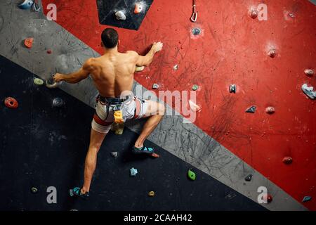 Panoramica del giovane uomo muscolare in allenamento indoor clymbing palestra. Vista posteriore di uno sportivo scalatore nello spostamento verso l'alto sulla roccia ripida, arrampicata su w artificiale Foto Stock