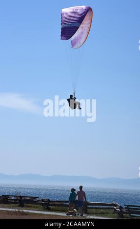 Victoria, British Columbia, Canada, 05 AGO 2020 - UN parapendio, approfittando del vento che esce dalle scogliere di Dallas Road e dal fronte oceano, sorvola i pedoni su un sentiero a Victoria sull'isola di Vancouver. Le montagne all'orizzonte sono le montagne olimpiche, nello Stato di Washington, Stati Uniti. Don Denton/Alamy Live News Foto Stock