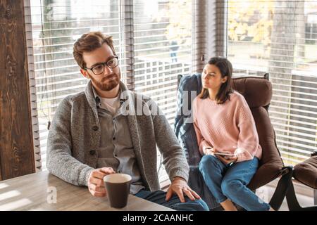 Giovane coppia attraente che discute l'acquisto di una nuova casa in una cucina moderna ben illuminata mentre parlano e bevono caffè, seduti sul Flo Foto Stock