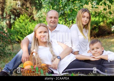 Soddisfare l'uomo e la donna dell'adulto che siede con la ragazza teen ed il ragazzo sul prato verde che ha picnic e sorridente alla macchina fotografica Foto Stock