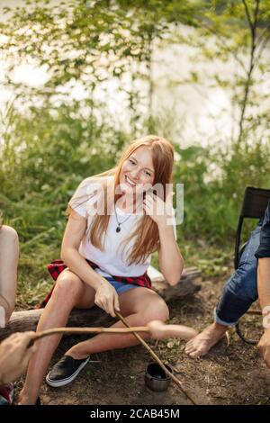 sorridente giovane zenzero che fissa i capelli e guardando la macchina fotografica mentre si siede a terra vicino al fuoco. vacanza, vacanza, giorno fuori, fine settimana Foto Stock