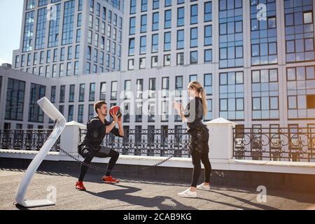 Bel ragazzo in giubbotto EMS che lancia una palla a una bella ragazza sul tetto dell'edificio moderno. Vista laterale foto. Studenti che si divertono all'aperto Foto Stock