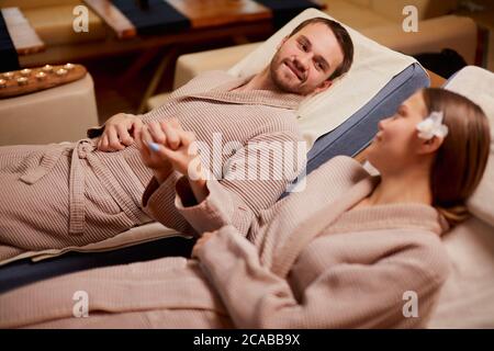 La coppia di sposi caucasici si stese su letti spa e si prende le mani l'una dell'altra, innamorati Foto Stock