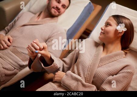 Carina coppia sposata caucasica sdraiata su letti spa e prendere le mani l'una dell'altra, innamorata. Indossando accappatoi Foto Stock