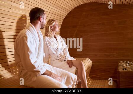 Foto laterale di giovani e bei amici seduti su una panca di legno caldo in un lussuoso hotel termale, cura del corpo, cura della pelle, benessere, concetto di benessere Foto Stock