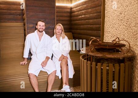 Bella giovane coppia passare il tempo felice nel centro di bellezza spa, seduta su panca di legno, indossando abiti da spogliatoio bianchi, guardando dritto alla macchina fotografica, it Foto Stock