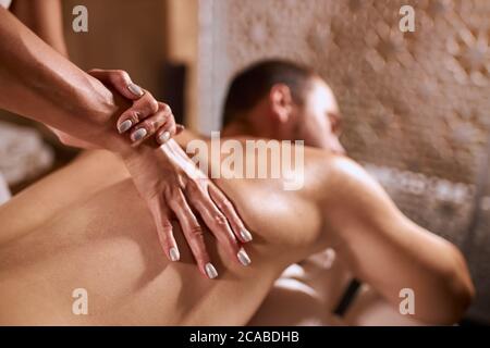 Vista ravvicinata delle mani delicate del massaggiatore asiatico professionale con una bella manicure, massaggiando la schiena del giovane uomo sdraiato sul tavolo da massaggio, sfondo sfocato Foto Stock