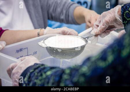 Le donne che si accovacciano le cagliate (versando / raccogliendo la cagliata morbida di formaggio di capra in contenitori o sacchetti di stoffa di formaggio durante una classe di cheesemaking in città di caduta, W. Foto Stock