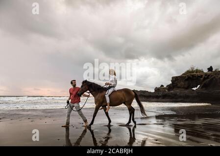 Asia, Indonesia, Bali, giovane caucasico con una bella giovane donna caucasica seduta a cavallo, su una spiaggia al tramonto, con tipica roccia balinese Foto Stock
