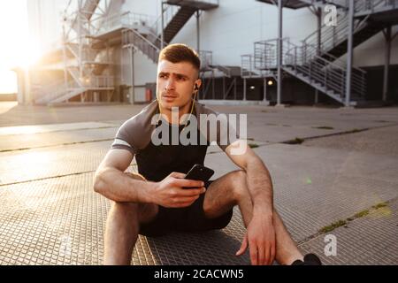 Immagine di un giovane sportivo caucasico che usa il cellulare e le cuffie mentre si siede sul pavimento all'aperto Foto Stock