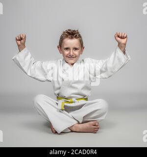 bambino allegro che pratica arti marziali seduto a terra con le braccia sollevate in un senso di vittoria. espressione felice. Foto Stock