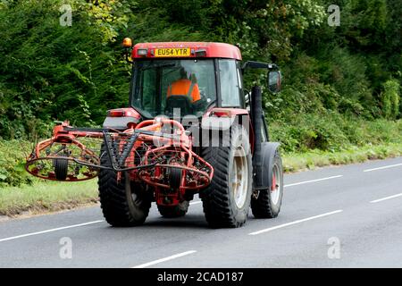 Un trattore con un rastrello da fieno, su una strada di campagna, Warwickshire, Regno Unito Foto Stock