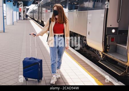 Donna in maschera protettiva in piedi sulla stazione ferroviaria vicino al treno con la sua valigia Foto Stock