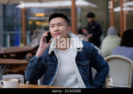 Primo piano un giovane asiatico seduto in un bar sul marciapiede, che effettua una telefonata. Sfocatura dello sfondo Foto Stock