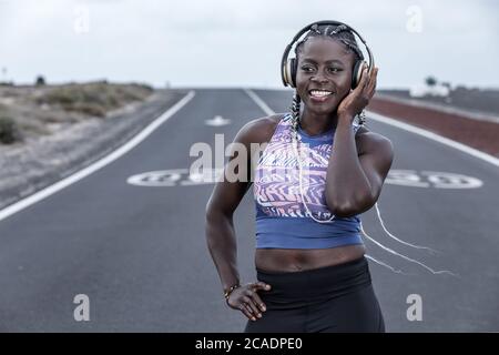 Donna sportiva nera positiva con una mano in vita sorridente e ascolto di musica in cuffia mentre si riposa sulla strada durante la sessione di corsa in periferia Foto Stock