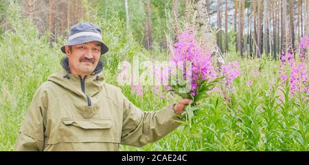 L'uomo anziano in tuta di zanzara tiene in mano bouquet di fireweed e guarda in macchina fotografica sullo sfondo di fiori prato e foresta Banner Chamaener Foto Stock